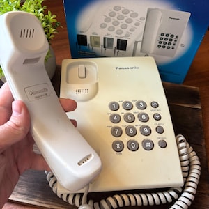 May include: Off-white Panasonic KX-TS2350UA corded telephone with a handset, number buttons, and a coiled cord. The phone is on a wooden surface, with the original box in the background.