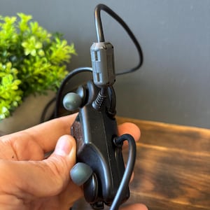 May include: A black electronic control module featuring two integrated earbud-style components with grey silicone tips. The device includes a thick black cable with a cylindrical ferrite core filter and a standard USB connector at the end of the cord.
