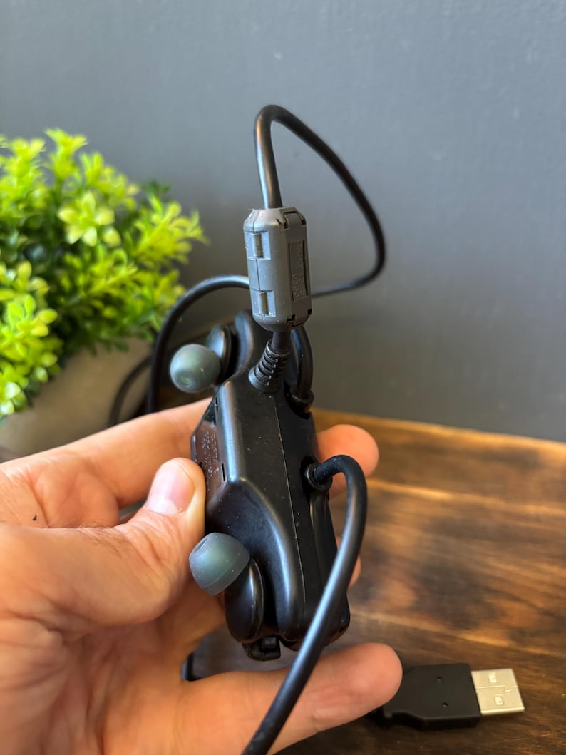 May include: A black electronic control module featuring two integrated earbud-style components with grey silicone tips. The device includes a thick black cable with a cylindrical ferrite core filter and a standard USB connector at the end of the cord.