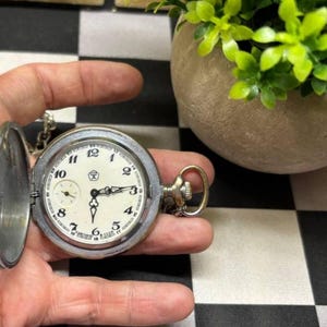 Op de afbeelding: Een zilverkleurig zakhorloge met een open scharnierend deksel, voorzien van een witte wijzerplaat met zwarte Arabische cijfers, sierlijke zwarte wijzers en een kleine sub-wijzerplaat. De wijzerplaat bevat een centraal logo en de tekst "Сделано в СССР".