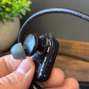 May include: A black wireless earbud with a translucent gray silicone tip and a connected black cable. The side of the earbud features playback control icons, a reset button, and the text RESET printed in white on the glossy plastic housing.