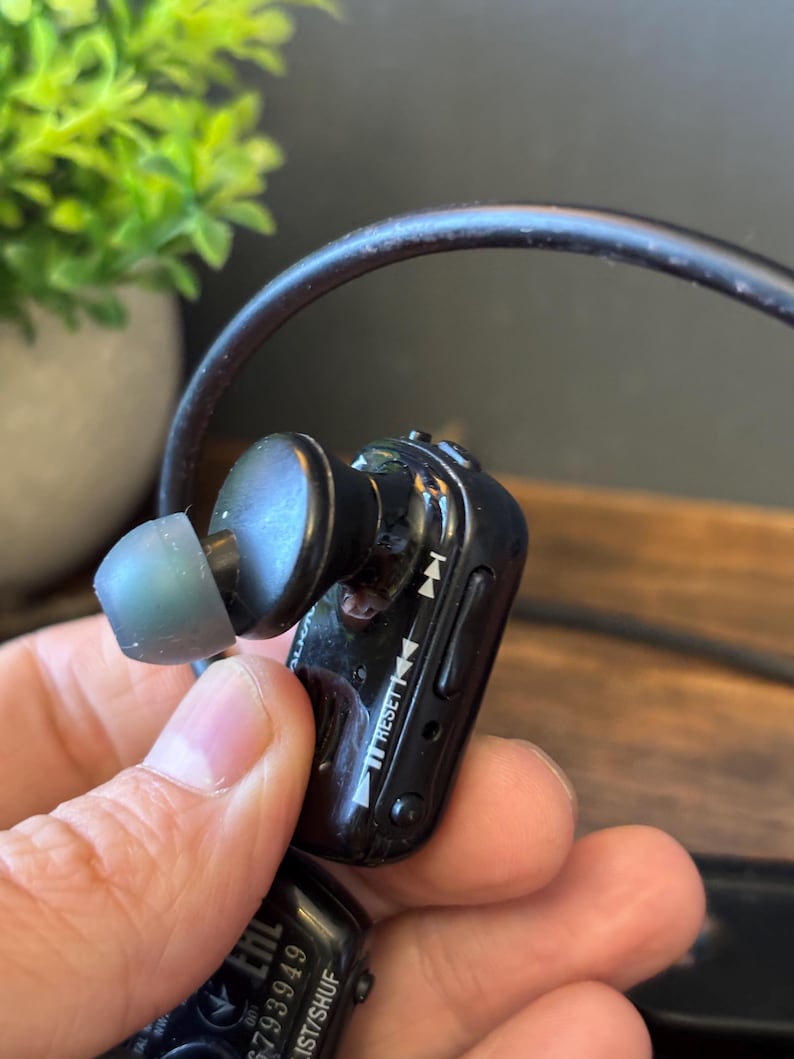May include: A black wireless earbud with a translucent gray silicone tip and a connected black cable. The side of the earbud features playback control icons, a reset button, and the text RESET printed in white on the glossy plastic housing.