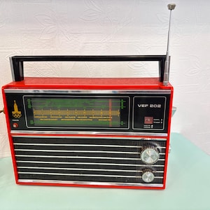 May include: A vintage red and black VEF 202 radio with a black handle and antenna. The radio has a green display and two silver knobs. The front of the radio has horizontal black lines. The radio is on a light green surface.