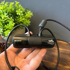 以下が含まれることがあります： 左側にSonyロゴ、右側にWalkmanロゴが配置された、水平な長方形デザインの黒いSony Walkmanデジタル音楽プレーヤー。このデバイスには、コネクタ付きの黒いケーブルと小さな円筒形の部品が取り付けられています。