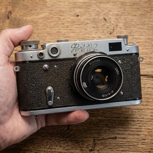 Puede incluir: Una cámara telemétrica vintage FED 2 con placa superior de metal plateado y cuerpo negro texturizado. El objetivo es negro con texto blanco 2.8/53 y la parte superior presenta grabados con el texto cirílico ФЭД 2 y el texto latino FED 2.