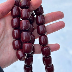 May include: A necklace of dark red, barrel-shaped beads. The beads are strung together with a white string, and the image is taken outdoors with a snowy background. The beads have a glossy finish.
