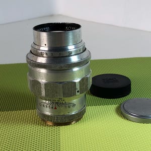 Può includere: Un obiettivo per fotocamera argentato con un tappo dell'obiettivo nero e un tappo dell'obiettivo argentato. L'obiettivo ha una scala di profondità di campo e un anello di apertura. L'obiettivo è in buone condizioni.