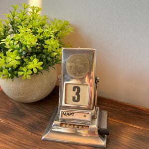 May include: A vintage silver-toned perpetual calendar displaying "3" on the date. The calendar has a decorative circular emblem with a building illustration. The word "MART" and "31" are visible, along with the word "СРЕДА". A small green plant in a white pot is in the background.