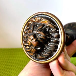 Puede incluir: Pomo de puerta de bronce con la cabeza de un león detallada dentro de un marco circular. La melena y los rasgos faciales del león están intrincadamente esculpidos. El pomo tiene una estética vintage, sugiriendo un estilo clásico.