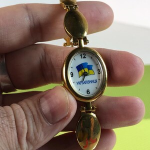 May include: A gold-tone wristwatch with a white face and a blue and yellow Ukrainian flag design. The watch face also has the text "УКРЗАЛІЗНИЦЯ" printed on it.