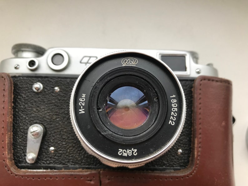 Może przedstawiać: Srebrno-czarny aparat fotograficzny 35 mm z brązowym sk&oacute;rzanym etui. Obiektyw aparatu ma nadrukowany tekst "W-26M" i "1896222" na boku. Obiektyw ma przysłonę 2.8/52.