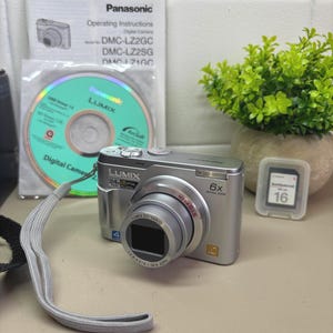 以下が含まれることがあります： 6倍光学ズームレンズを搭載したシルバーのパナソニックLUMIXデジタルカメラ。カメラにはCD、取扱説明書、16GBのメモリーカードが付属しています。カメラレンズにはLUMIX DC VARIOと記載されています。