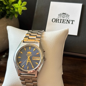 May include: An Orient wristwatch with a silver-tone stainless steel link bracelet and a round case. The watch features a dark blue dial with gold-tone hour markers and hands, a day-date window at the three o'clock position, and the Orient logo.