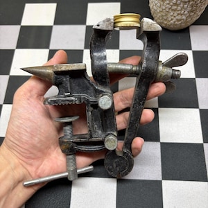 May include: A small, antique black metal vise with an integrated anvil. The vise is holding a gold-colored metal object. The tool has a screw mechanism and a handle for tightening. The background is a black and white checkered pattern.