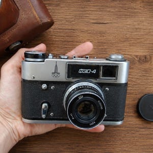 May include: A vintage ФЭД-4 camera with a silver and black body, held in a hand. The camera has a lens, dials, and the text "ФЭД-4". A brown leather case is visible in the background, along with a lens cap.