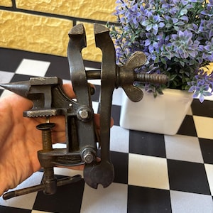 以下が含まれることがあります： アンティークスタイルの小さな黒い金属製の万力。金床、ネジ、蝶ナットが付いています。万力は手で持たれており、背景には白と黒のチェック柄の表面があります。紫色の花の小さなポットも背景にあります。