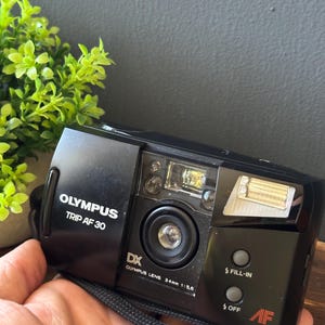 May include: A black Olympus Trip AF 30 point-and-shoot film camera with a 34mm 1:5.6 lens. The front features the Olympus logo, DX branding, a flash unit, and buttons labeled FILL-IN and OFF, along with a black wrist strap attached to the side.