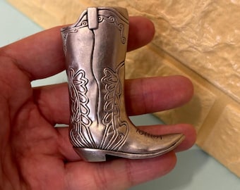 May include: A silver-colored cowboy boot figurine with intricate embossed floral designs. The boot has a pointed toe and a small heel. The metal has a slightly aged appearance, adding to its vintage charm. The boot is held in a hand.