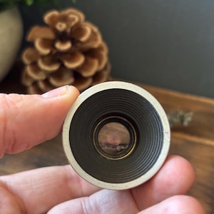 Puede incluir: Un accesorio de lente de cámara circular con un borde exterior de metal plateado y un barril interior negro con finas ranuras concéntricas. El centro de la lente revela un elemento de vidrio con un revestimiento reflectante y texto.