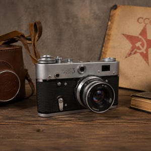 May include: A silver and black FED 3 rangefinder camera with a lens, featuring a brown leather carrying case to the left and a tan book with a red star and the text CCCP printed on the cover positioned to the right of the camera body.