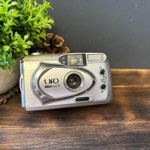 May include: A silver UFO Mini Lux II compact film camera featuring a central lens, a built-in flash unit, and three small black control buttons on the right side. The camera body has a metallic finish with black text branding on the front.