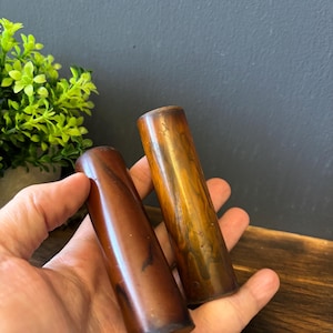 Op de afbeelding: Twee cilindervormige objecten met een gladde, gepolijste afwerking. De ene cilinder heeft een diepe, donkerroodbruine tint met subtiele donkere strepen, terwijl de andere een lichtere, doorschijnende amberbruine kleur met wervelende goudbruine patronen vertoont.