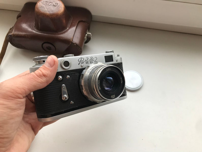 Może przedstawiać: Srebrny i czarny aparat fotograficzny 35 mm z obiektywem oznaczonym "Industar 50-2". Aparat ma czarny sk&oacute;rzany futerał.