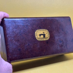 Op de afbeelding: Een vintage bruine naaimachinetas met een goud en zwart embleem. Het embleem luidt "VEB Nähmaschinenwerk Saalfeld/Saale" en toont een silhouet van een naaimachine. De rechthoekige tas lijkt van hout te zijn.
