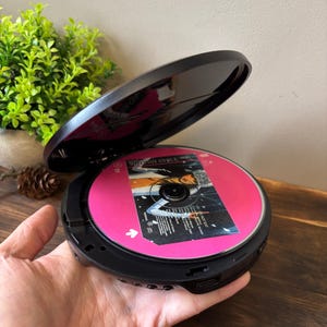 以下が含まれることがあります： ピンク色のCDが入った黒色のポータブルCDプレーヤー。CDには「Winter Chill」の文字が書かれています。プレーヤーは開いており、CDが見えています。プレーヤーは手で持たれています。
