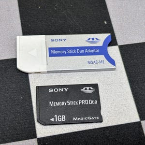 Peut inclure: Un adaptateur Sony Memory Stick Duo et une carte Memory Stick PRO Duo de 1 Go. L'adaptateur est argenté et blanc avec des accents bleus, et la carte est noire. Les deux articles portent le logo Sony et des informations.