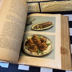 Op de afbeelding: Open vintage kookboek met twee voedselillustraties. Eén toont een lang, rechthoekig gerecht met een bruin, gehaktbroodachtig item op kruiden. De andere toont zes ronde, goudbruine voedselitems op een zilveren bord.