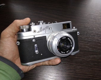 Może przedstawiać: Srebrno-czarny aparat fotograficzny 35 mm z czarnym skórzanym pokryciem. Aparat ma obiektyw oznaczony jako "Industar-50" i numer seryjny "N5915097". Aparat ma podpis, który brzmi "Myro" na górze.