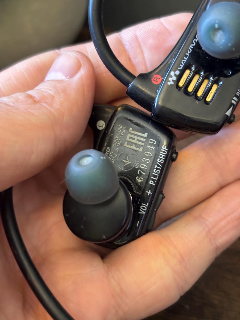 May include: Black Sony Walkman wireless in-ear headphones with a wrap-around neckband. The device features grey silicone ear tips, volume and playlist control buttons, and printed text including the serial number 6793949 and Made in Malaysia.