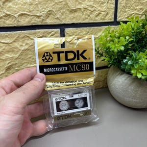 May include: A TDK Microcassette MC90 in its original packaging. The gold and black package displays the TDK logo and product details. The cassette is visible through clear plastic, with the text "Ideal for dictation and telephone answering machines."
