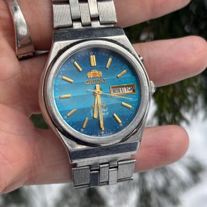 May include: A vintage Orient watch with a silver-tone metal band and a blue dial. The watch face features gold-tone hour markers and hands, with a day and date display. The watch is a classic timepiece.
