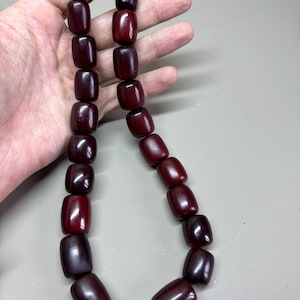 Puede incluir: Un collar de cuentas rojas oscuras en forma de barril. Las cuentas están ensartadas, formando un collar. Las cuentas tienen un acabado brillante y varían ligeramente en color, desde el burdeos oscuro hasta un rojo más claro. El collar está sostenido por una mano.