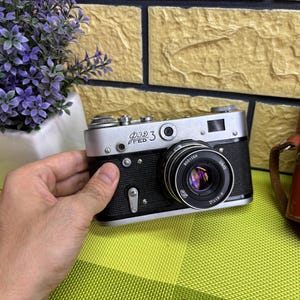 May include: Silver and black FED 3 vintage camera with a lens and a leather case. The camera is held in a hand, with a purple flower arrangement in the background.