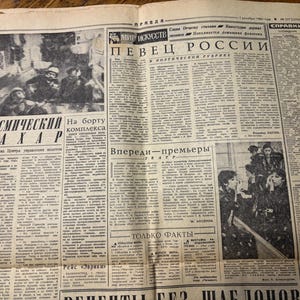 May include: A black and white newspaper clipping with the headline "Kosmicheskiy Sakhar" and a photo of a group of people in space suits. The article discusses the "Kosmicheskiy Sakhar" project and the crew on board the complex. The newspaper also features a section titled "Vpered - Premiery" and another section titled "Tol'ko Fakty".