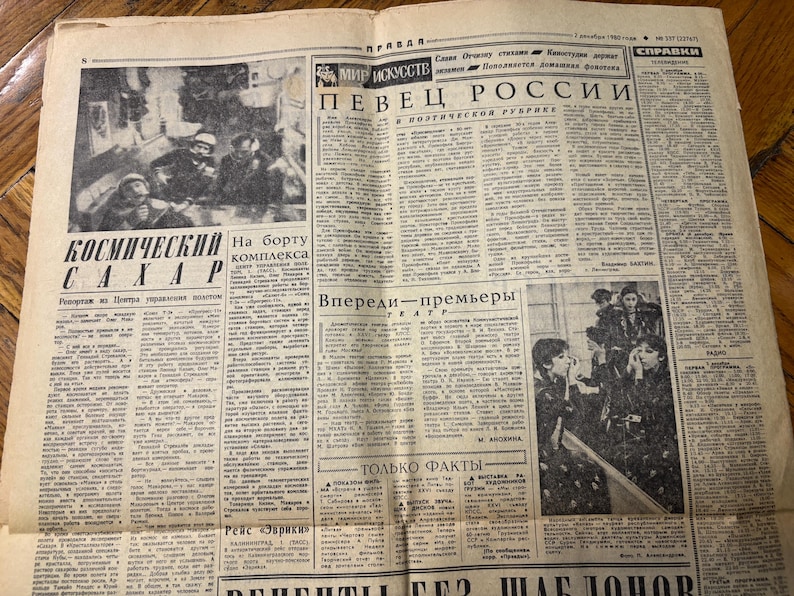 May include: A black and white newspaper clipping with the headline "Kosmicheskiy Sakhar" and a photo of a group of people in space suits. The article discusses the "Kosmicheskiy Sakhar" project and the crew on board the complex. The newspaper also features a section titled "Vpered - Premiery" and another section titled "Tol'ko Fakty".