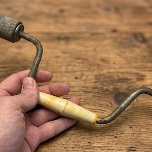 May include: A vintage hand brace drill featuring a metallic crank handle, a central cream-coloured cylindrical grip, and a black rotating knob at the end. The tool includes a metal chuck at the top holding a small drill bit for manual drilling.