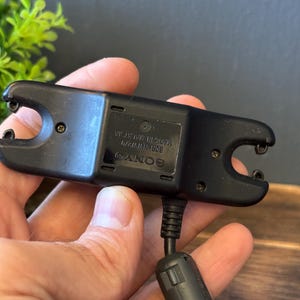 May include: A black Sony battery charger with a rectangular body, two U-shaped contact ends, and a power cable featuring a cylindrical ferrite bead. The device is labeled with the text SONY, BCR-NW870, and MADE IN MALAYSIA on the center panel.