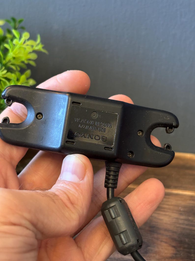 May include: A black Sony battery charger with a rectangular body, two U-shaped contact ends, and a power cable featuring a cylindrical ferrite bead. The device is labeled with the text SONY, BCR-NW870, and MADE IN MALAYSIA on the center panel.