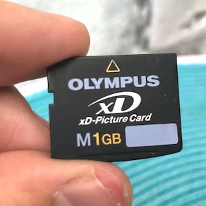 May include: Black Olympus xD-Picture Card, held between fingers. The card has white text that reads "OLYMPUS", "xD-Picture Card", and "M1GB". A yellow triangle is at the top.