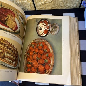 Op de afbeelding: Pagina uit een vintage kookboek met voedselillustraties. Eén afbeelding toont een cake met rode saus op een wit bord. Een andere toont een taart met een decoratieve korst. Een kom met aardbeien en room is ook zichtbaar.