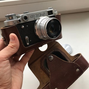 Może przedstawiać: Srebrny aparat fotograficzny w brązowym sk&oacute;rzanym etui. Aparat ma obiektyw z napisem "FED-2".