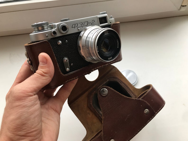 Może przedstawiać: Srebrny aparat fotograficzny w brązowym sk&oacute;rzanym etui. Aparat ma obiektyw z napisem "FED-2".