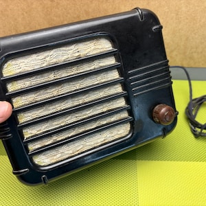 May include: A vintage black radio with a cloth speaker grille and a brown knob. The radio has a cord attached to it.