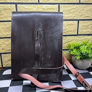 May include: A dark brown leather messenger bag with a pink canvas strap. The bag has a rectangular shape with a flap closure and a stitched detail down the center. The strap is adjustable with metal hardware.