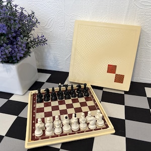 Op de afbeelding: Een vintage schaakspel met zwarte en witte stukken op een rood-wit geblokt bord. De set bevat een crèmekleurige koffer met een geblokt patroon en twee rode vierkanten. Een kleine pot met paarse bloemen is ook aanwezig.