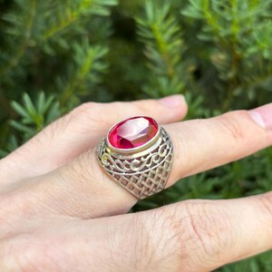 May include: A silver ring featuring a large, oval-cut, vibrant red gemstone set in a raised bezel. The band displays an intricate, openwork lattice design that extends from the base of the setting down the sides of the metal shank.